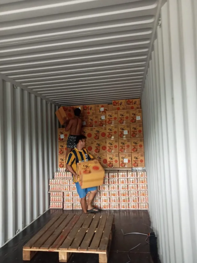 Workers hand-stack cup-noodle cartons above rows of canned drinks inside the container.