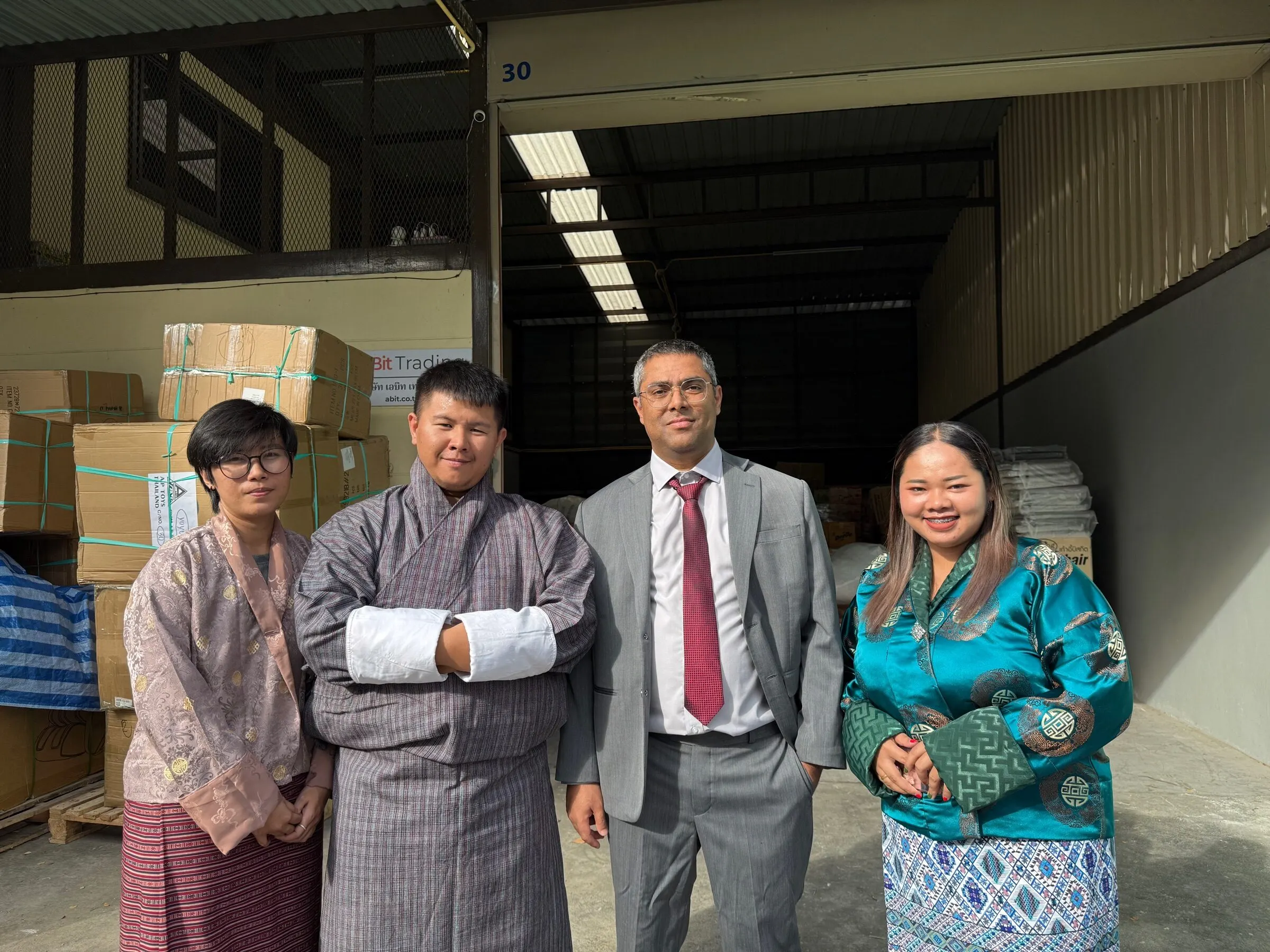 The aBit Trading team — three of them in Bhutanese gho and kira — standing at the doors of the company's Bangkok warehouse