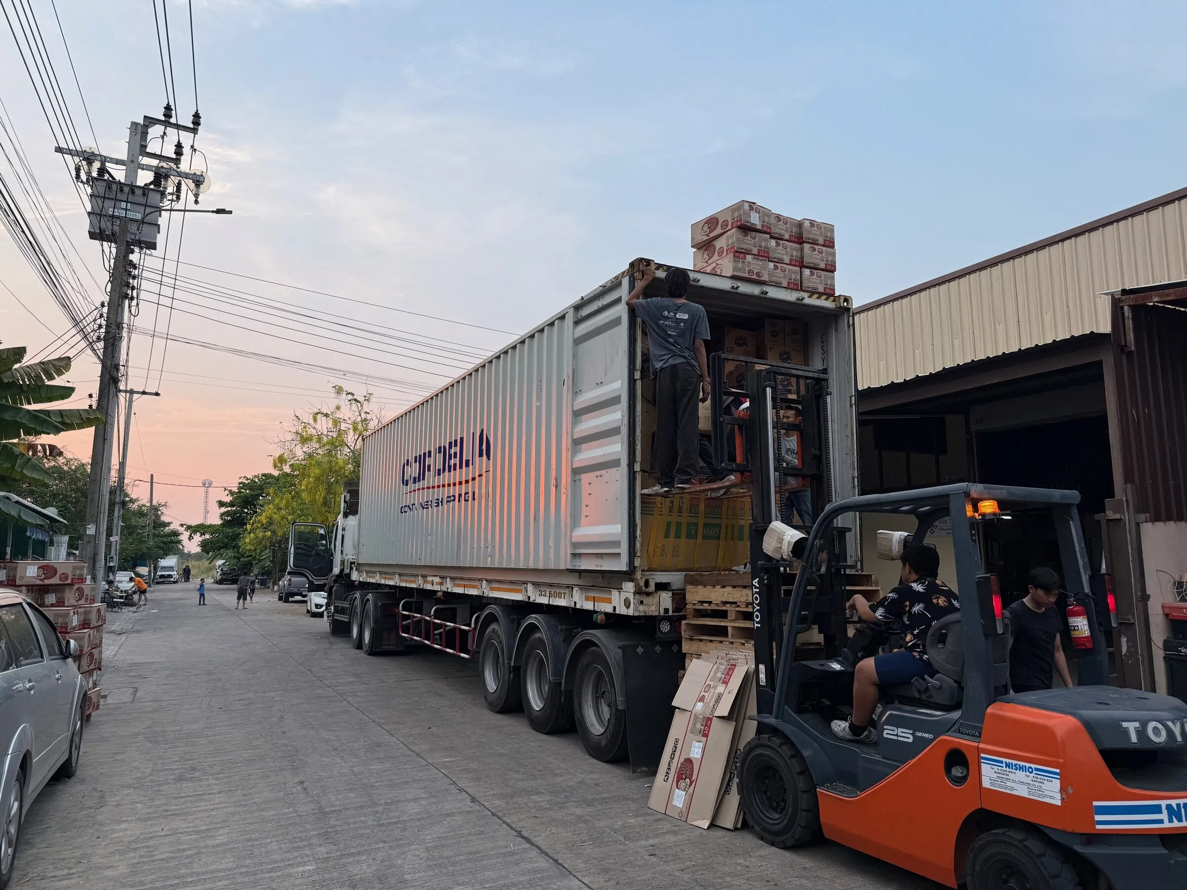 Container loading continuing at the Bangkok warehouse at dusk, with cartons stacked at the doors